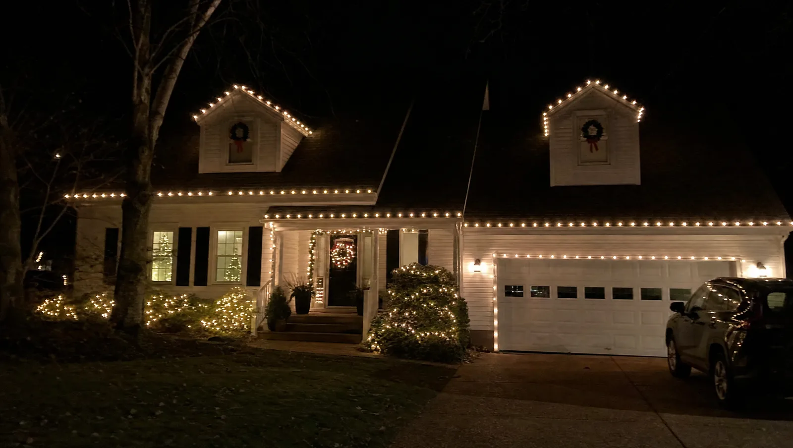 A white cape-style home at night with warm-white bulbs tracing every roofline and dormer, lit wreaths in the upstairs windows, and lit shrubs along the front beds