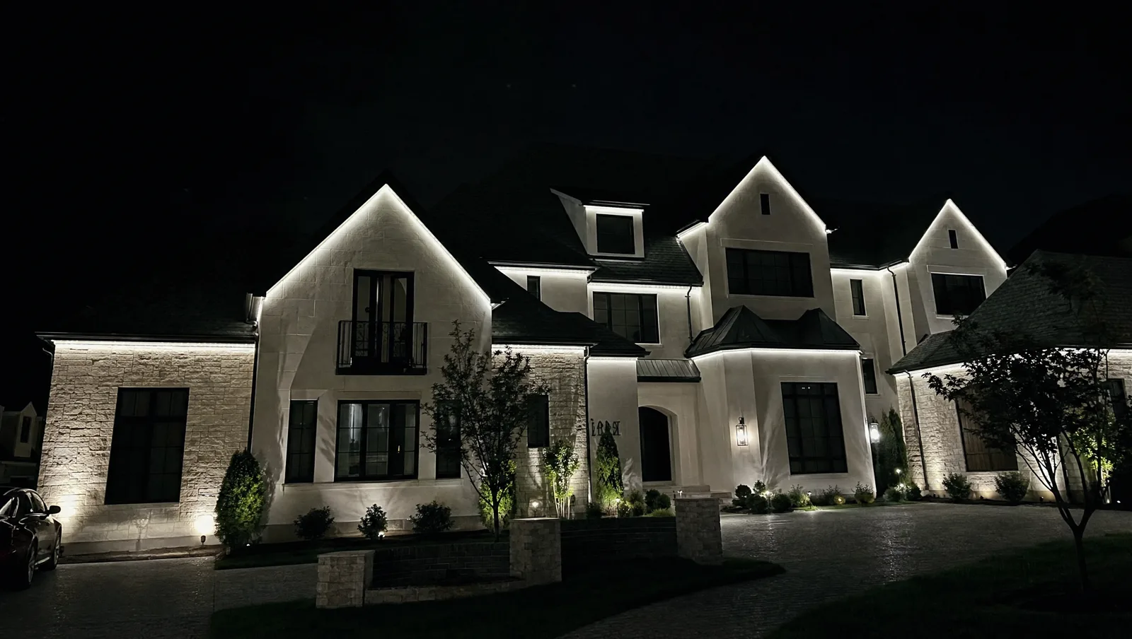 A large stone home at night with warm-white light washing down the gables, eaves and stonework from lighting integrated into the roofline